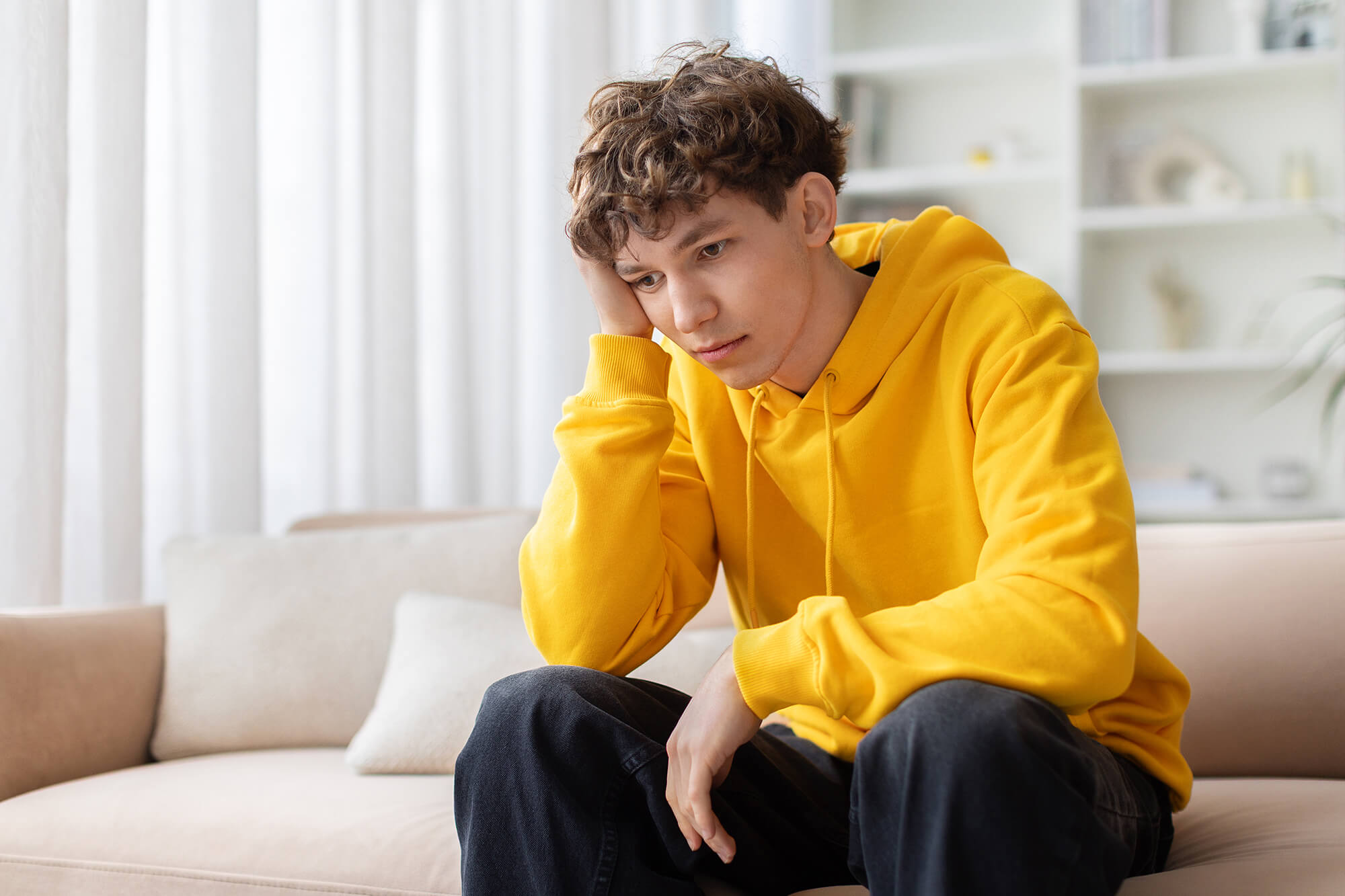 A teen in a yellow hoodie sitting on a couch, looking down